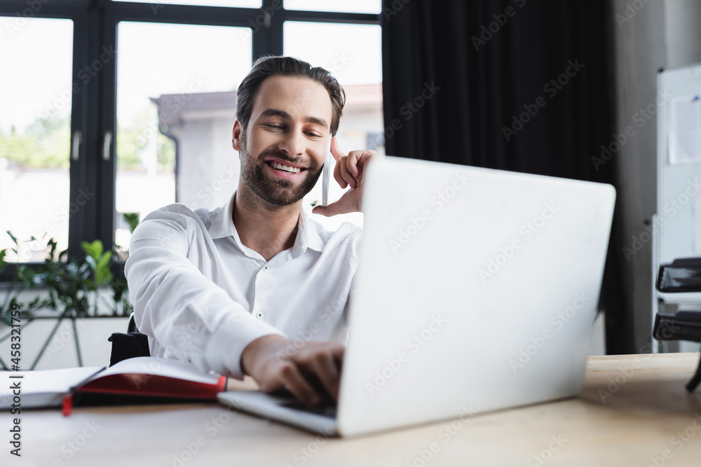 happy businessman using blurred laptop while talking on smartphone in office