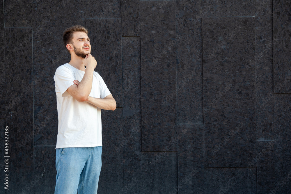 Emotions, gesture, expression and people concept - A pondering Caucasian bearded man in a white T-shirt and jeans looking at the copy space on the right side outside on a black background Emotions, gesture, expression and people concept - A pondering Caucasian bearded man in a white T-shirt and jeans looking at the copy space on the right side outside on a black background