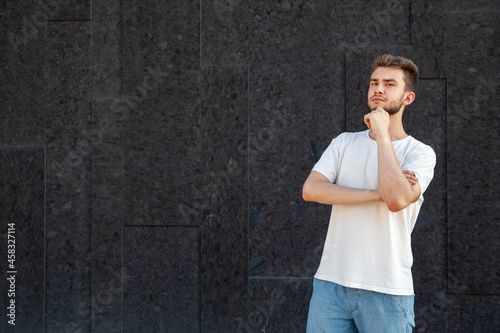 Emotions, gesture, expression and people concept - A thinking Caucasian bearded man in a white T-shirt and jeans looking into a camera with copy space on the left side outside on a dark background