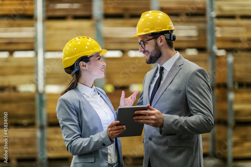 Warehouse visiting. Business people wear suit a protective yellow helmet stand in the warehouse. They hold the tablet in their hand making eye contact. Business conversation in a work environment