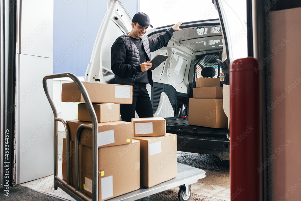 Courier in uniform holding a digital tablet checking amount of packages on a cart while standing at a car Courier in uniform holding a digital tablet checking amount of packages on a cart while standing at a car