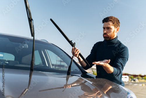 Caucasian man feeling confused while changing windscreen wipers on a car at the street during the sunny day. Transportation concept