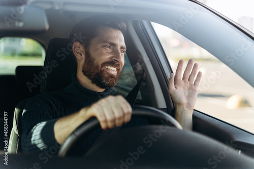 Handsome young smiling driver of car waving hand as a sign of greeting while driving the car with pleasure emotions. Transportation concept