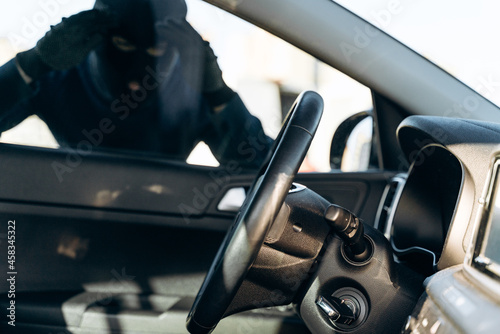 фотографія View from the car at the man dressed in black with a balaclava on his head looking at the glass of car before the stealing