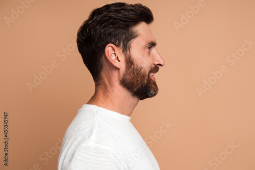 Side view of positive handsome brunette man with beard in white shirt standing with pleasure smile and looking away with joyful expression. Indoor studio shot isolated on beige background