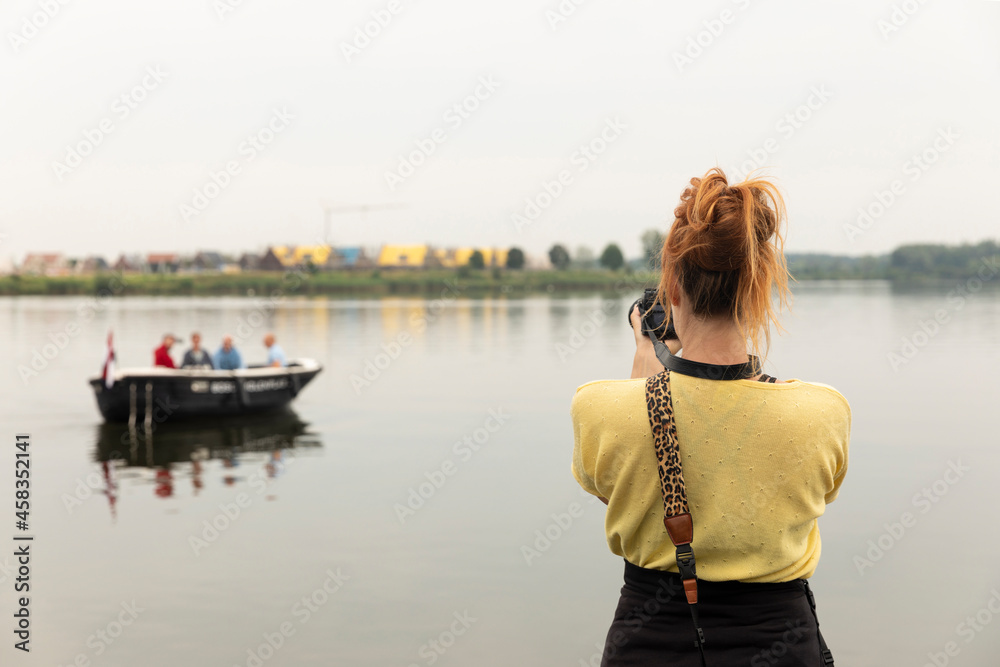 Female tourist photographer taking pictures of a small boat with people in it in Thorn, the Netherlands surrounded by the water in the river. Capturing the holiday, creating memories