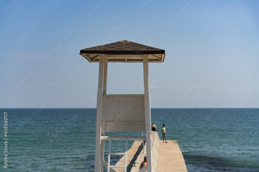 Behind the wooden observation tower on the beach, a concrete pier goes deep into the sea on a sunny day against the background of blue sky and sea