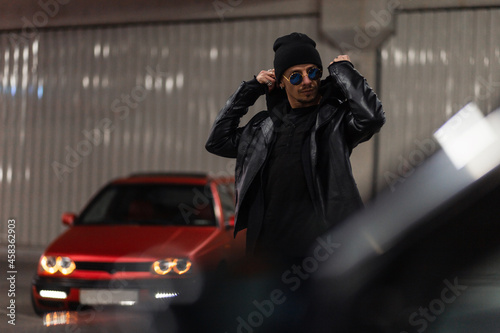 Stylish young handsome man with sunglasses in a fashionable leather jacket with a stylish hoodie puts on a hood in the night city at the parking