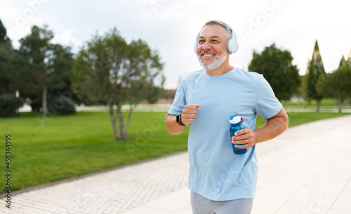 Happy mature man wearing headphones with water bottle in hand jogging outside in city park