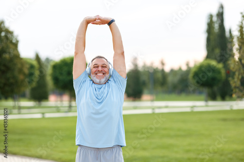 Active senior man in cordless headphones doing body stretching with hands up outside in city park