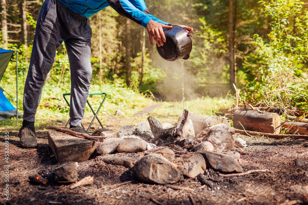 Man extinguishing campfire with water from cauldron in summer forest. Put out campfire. Traveling fire safety rules - obrazy, fototapety, plakaty 