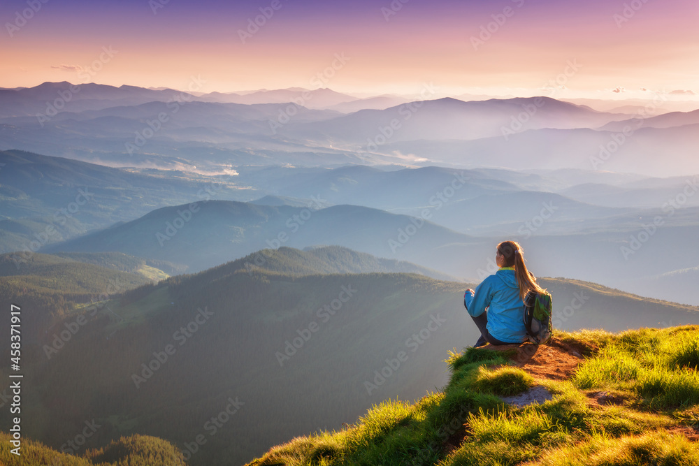 Young woman with backpack sitting on the mountain peak and beautiful mountains in fog at sunset in autumn. Landscape with sporty girl, green grass, forest, hills, sky, sunbeams in fall. Travel. Nature