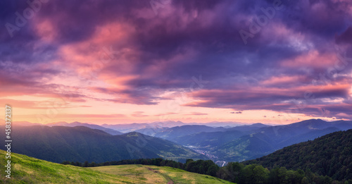 Mountains at beautiful sunset in summer. Colorful panoramic landscape with meadows with green grass, sky with vibrant clouds, mountains with forest. Trail on the hill. Travel and nature. Scenery