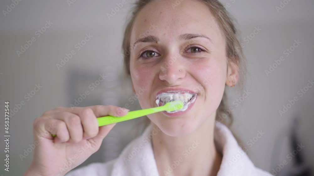 Close-up of brunette young woman with brown eyes brushing teeth in slow motion smiling looking at camera. Portrait of confident slim beautiful Caucasian lady taking care of dental health at home Close-up of brunette young woman with brown eyes brushing teeth in slow motion smiling looking at camera. Portrait of confident slim beautiful Caucasian lady taking care of dental health at home