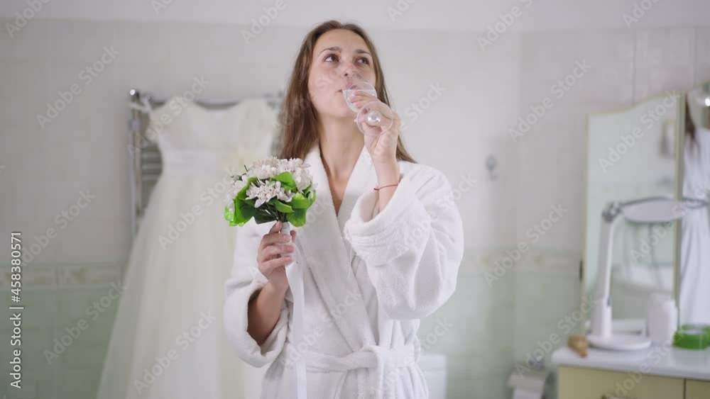 Front view of smiling charming young Caucasian bride drinking champagne smelling bouquet of flowers in the morning on wedding day. Portrait of happy confident woman in bathrobe at home indoors Front view of smiling charming young Caucasian bride drinking champagne smelling bouquet of flowers in the morning on wedding day. Portrait of happy confident woman in bathrobe at home indoors