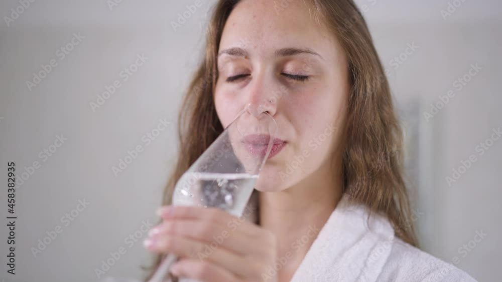 Close-up of happy smiling young woman with bridal bouquet drinking champagne standing in bathroom with blurred dress at background. Portrait of confident charming slim bride at home indoors