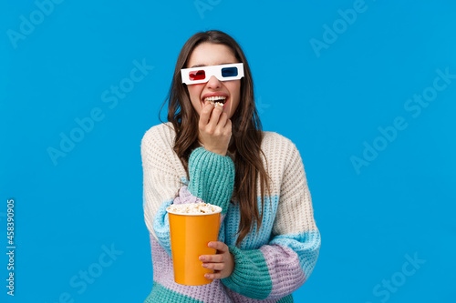 Cheerful and happy, laughing brunette caucasian girl enjoying funny movie, wearing 3d glasses, chuckling and munching popcorn, holding box, standing in winter sweater, cinema offer student dicsount photo