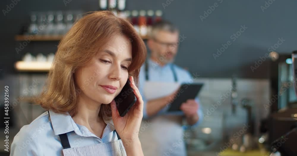 Close up portrait of beautiful happy woman in apron speaking on smartphone at workplace in cafe while male colleague typing on tablet on background. Family business, coffee house, job concept Close up portrait of beautiful happy woman in apron speaking on smartphone at workplace in cafe while male colleague typing on tablet on background. Family business, coffee house, job concept