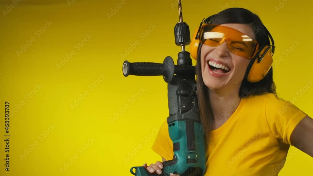 A cheerful woman in protective headphones is holding a drill in her hand