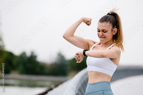 Woman in sports clothes, looking at her smart watch and showing her arm muscles. With a winning and celebration expression. Women, fitness, psychology, personal growth and sport concept. Copyspace