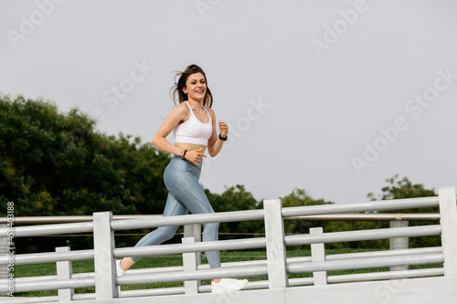 Woman running, wearing sports clothes. Smiling. With a bridge and park in the background. Women, fitness, psychology, personal growth and sport concept. Copyspace