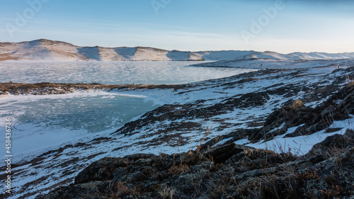 Two frozen lakes are separated by a thin strip of land. Snow lies on the ice and on the hillsides. In the foreground are boulders and dry grass. Blue sky. A sunny winter day. Baikal