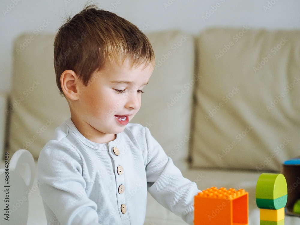 Naklejka premium Cute toddler boy in beige clothes playing blocks in bright interioir