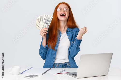 Relieved and happy girl becomes rich after making great deal, shaking hands, fist pump saying yes and raising head up sky delighted, holding big money, cash near dest with laptop, white background photo