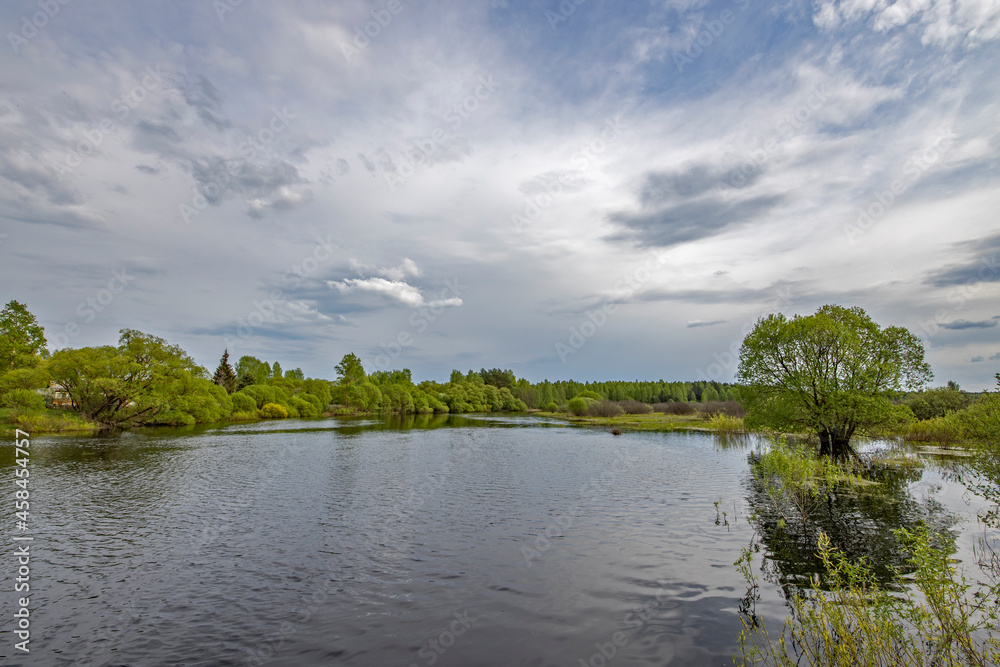 Landscape with a dramatic sky reflected in the river. Early spring, juicy May greens. Bright green foliage on trees and bushes. Landscape with a dramatic sky reflected in the river. Early spring, juicy May greens. Bright green foliage on trees and bushes.
