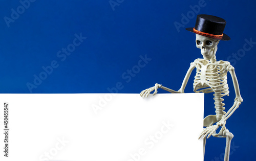Human skeleton in black hat holds blank sing board on blue background. Zombie behind white banner for advertisement with copy space. Concept of coming soon of Halloween, discounts, sales.