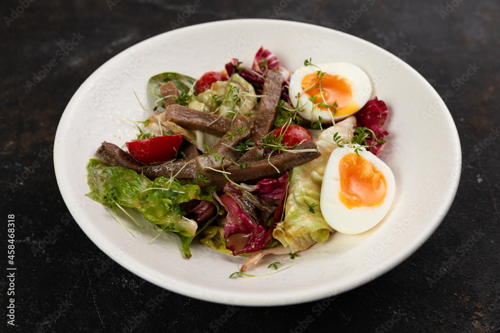 A beautiful Fresh salad with meat tongue vegetables lettuce leaves egg cucumbers bell pepper Cherry tomatoes olives red onions is decorated on a white plate on a black background large serving A beautiful Fresh salad with meat tongue vegetables lettuce leaves egg cucumbers bell pepper Cherry tomatoes olives red onions is decorated on a white plate on a black background large serving