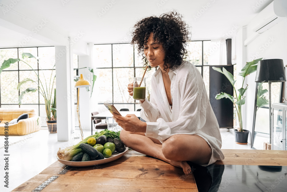 young mixed race woman looking at cellphone having green juice breakfast. sitting on kitchen counter.