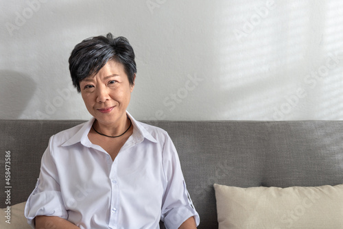 Close-up portrait of an attractive middle-aged mature grey-haired Asian woman posing at home in the morning, elder female sitting on a sofa in the living room. Positive single senior retired Concept