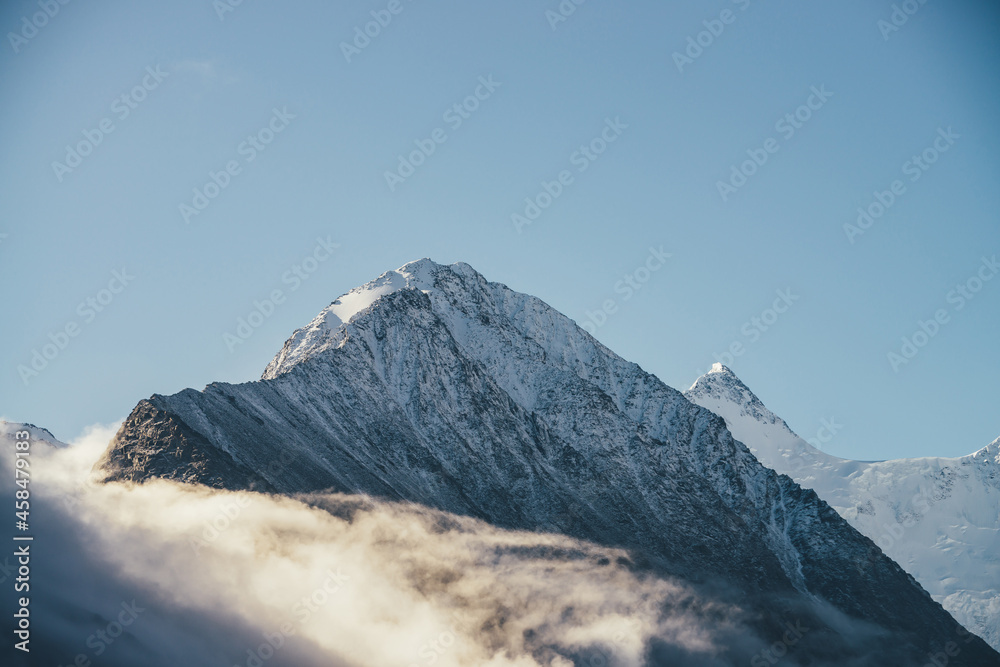 Fototapeta premium Beautiful view of snow-capped mountains above thick clouds in sunshine. Scenic bright mountain landscape with white-snow peak among dense low clouds in blue sky. Wonderful scenery with snowy pinnacle.