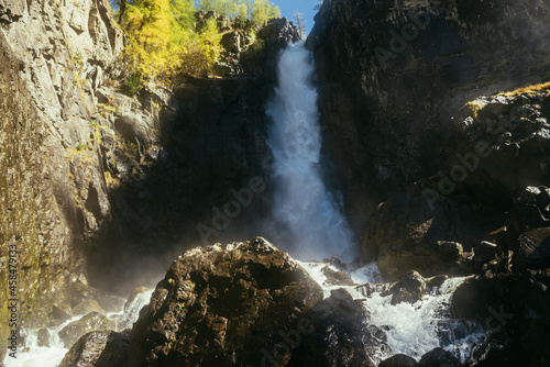 Scenic autumn landscape with vertical big waterfall and yellow trees at mountain top in sunshine. Powerful large waterfall in rocky gorge. High falling water and trees of golden colors in fall time.