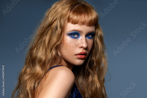 An attractive beautiful woman with natural curly shiny hair poses. A lot of hairstyle with the effect of beach curls. Red-haired high fashion model with freckles on her face with blue evening make-up. © strekozza77