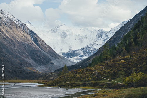 Scenic alpine landscape with mountain lake in valley in autumn colors with view to beautiful high snowy mountains in sunshine. Awesome scenery with autumn valley and great snow-white mountain wall.