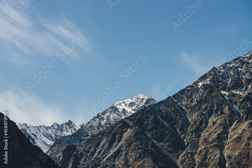 Alpine landscape with high snowy mountain with peaked top under cirrus clouds in sky. Atmospheric view to big snow covered mountains in sunshine. Black rocks and white-snow pointy peak in sunlight. © Daniil