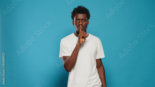 Young man doing silence gesture with finger on mouth in studio. Confident person asking for secrecy and quiet, using forefinger to make censorship expression over isolated background.