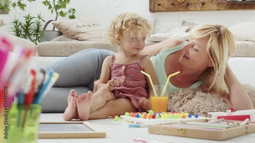 smiling mother smiles at curly haired blonde daughter child, drinking cheerfully from the same glass dividing orange juice with straws, preschool learning activity at home, concept of healthy growing