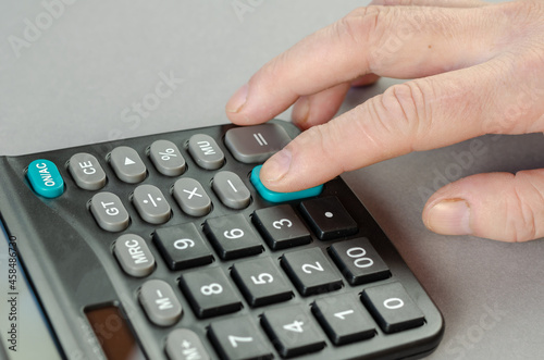 The man's index finger presses the green plus key of the calculator. The businessman performs mathematical calculations on the electronic device. Indoors. Selective focus.
