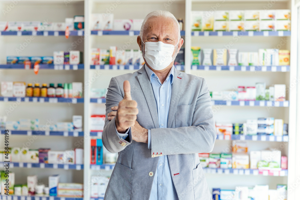 Portrait of a male pharmacist showing a sign of approval with his hand, thumb up. A man with gray hair in a modern uniform and gesturing approval with one hand. Look at the camera Protective face mask