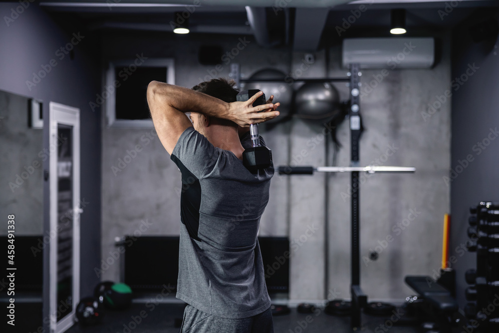 The man keeps good physical shape and fitness. Lifting dumbbells between the shoulder blades in a closed gym with a gray interior. Sports routine and healthy lifestyle The man keeps good physical shape and fitness. Lifting dumbbells between the shoulder blades in a closed gym with a gray interior. Sports routine and healthy lifestyle