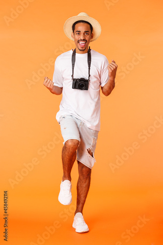 Full-length vertical shot excited good-looking african american guy on joyrney abroad, travelling feeling amused, fist pump, winning trip travel agency, dancing, standing orange background photo