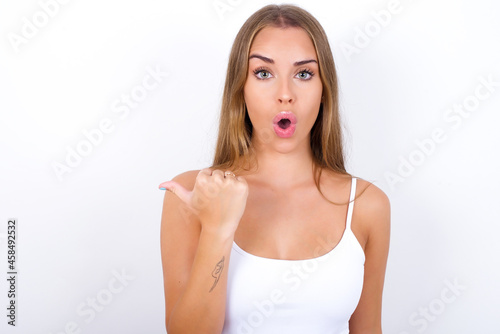 Shocked beautiful blonde girl wearing sexy t-shirt on white background points with thumb away, indicates something. Check this out. Advertisement concept.