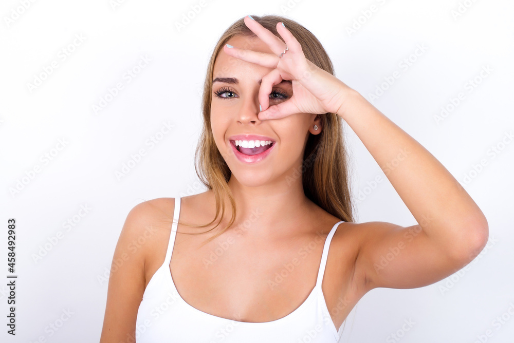 beautiful blonde girl wearing sexy t-shirt on white background with happy face smiling doing ok sign with hand on eye looking through finger. beautiful blonde girl wearing sexy t-shirt on white background with happy face smiling doing ok sign with hand on eye looking through finger.