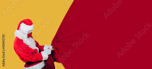 modern santa claus with red face mask pulls with all his might a giant christmas sack - banner copy space for writing and graphics