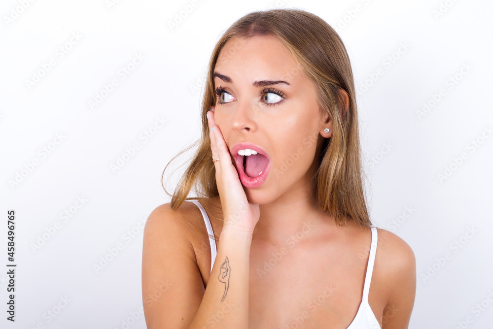 beautiful blonde girl wearing sexy t-shirt on white background excited looking to the side hand on face. Advertisement and amazement concept. beautiful blonde girl wearing sexy t-shirt on white background excited looking to the side hand on face. Advertisement and amazement concept.