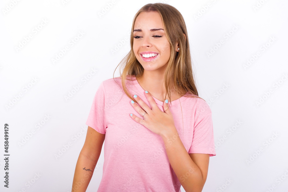 Joyful beautiful blonde girl wearing pink t-shirt on white background expresses positive emotions recalls something funny keeps hand on chest and giggles happily. Joyful beautiful blonde girl wearing pink t-shirt on white background expresses positive emotions recalls something funny keeps hand on chest and giggles happily.