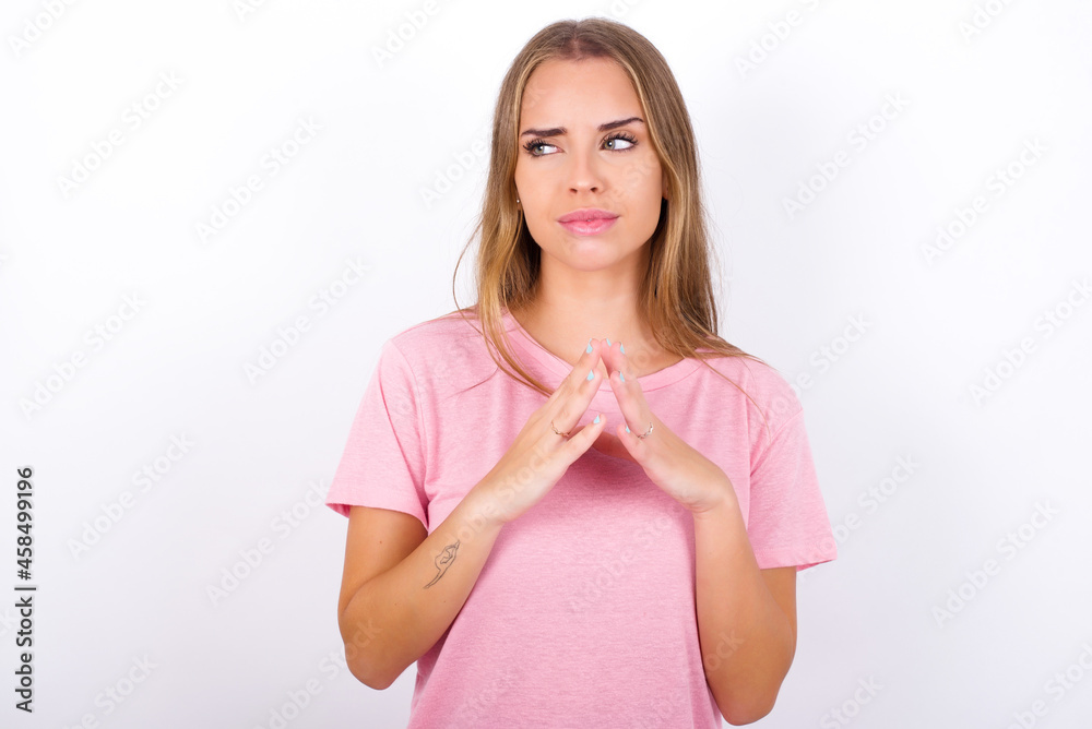 beautiful blonde girl wearing pink t-shirt on white background steepls fingers and looks mysterious aside has great evil plan in mind beautiful blonde girl wearing pink t-shirt on white background steepls fingers and looks mysterious aside has great evil plan in mind